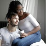 Positive young multiracial couple in casual clothes sitting on comfortable sofa with cup and looking away through window in modern living room