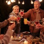 Laughing friends with kids spending time in backyard at night enjoying dinner with garlands and clinking glasses