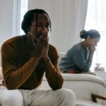 Wistful concerned African American couple in casual clothing sitting on bed at home after having quarrel